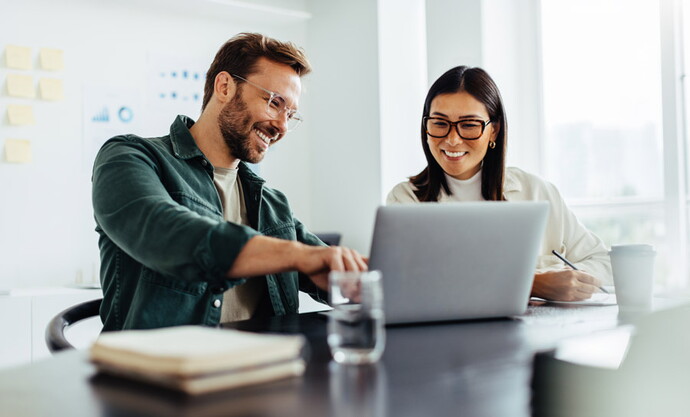 Vier junge Menschen diskutieren lachend in einem modernen Büro bei Nachtbeleuchtung, zwei sitzen am Tisch mit Laptop, einer hält ein Tablet