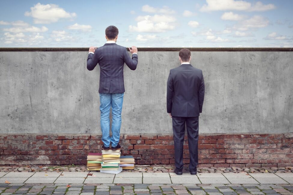 Two men by a wall, one standing on books, symbolizing knowledge advantage.