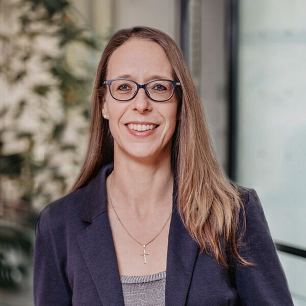 Woman with glasses and a cross necklace in business attire against a bright background