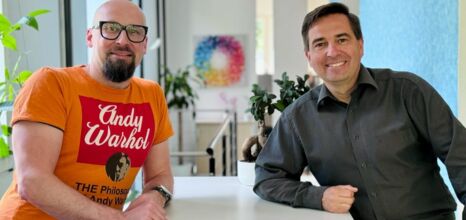 Ronny Büchner and Tino Favetto sit next to each other and smile into the camera.