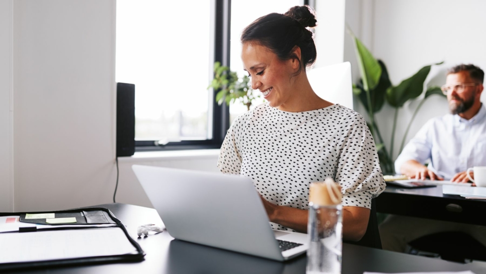 Smiling woman working on a laptop in a modern office, with a colleague in the background.