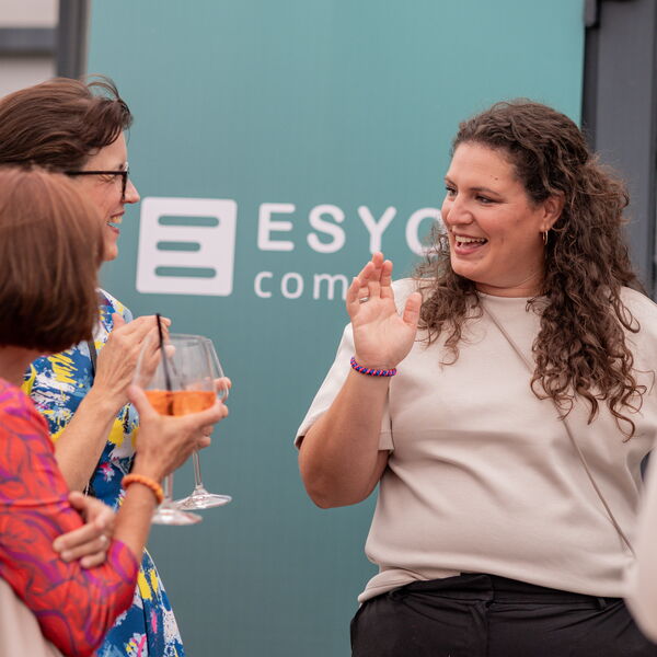Zwei Frauen unterhalten sich lachend vor einer türkisen Wand mit ESYON-Logo, eine hält ein Glas mit orangefarbenem Getränk