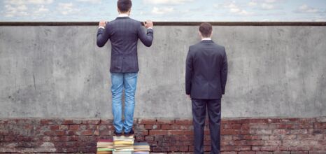 Two men by a wall, one standing on books, symbolizing knowledge advantage.